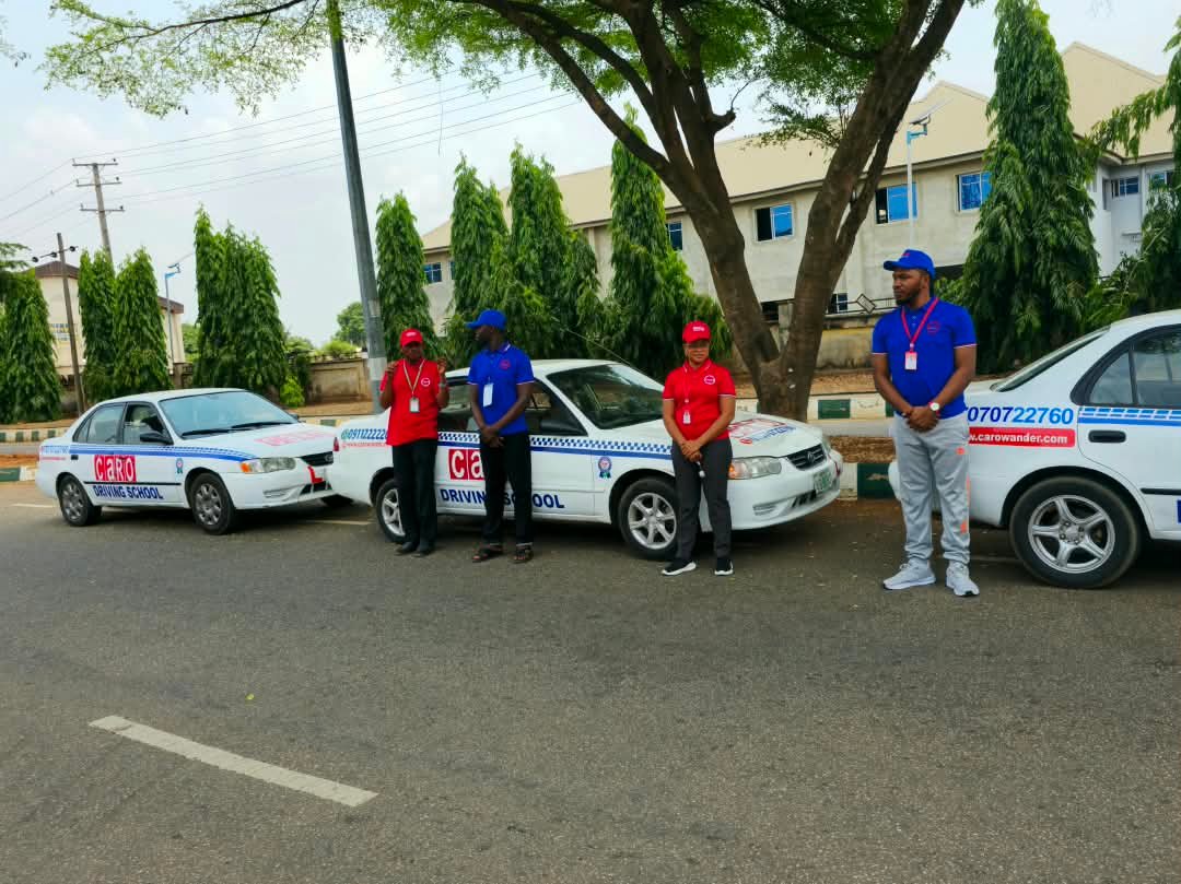 Four CARO Wander staff in red and blue uniforms beside three white CARO Driving School training cars